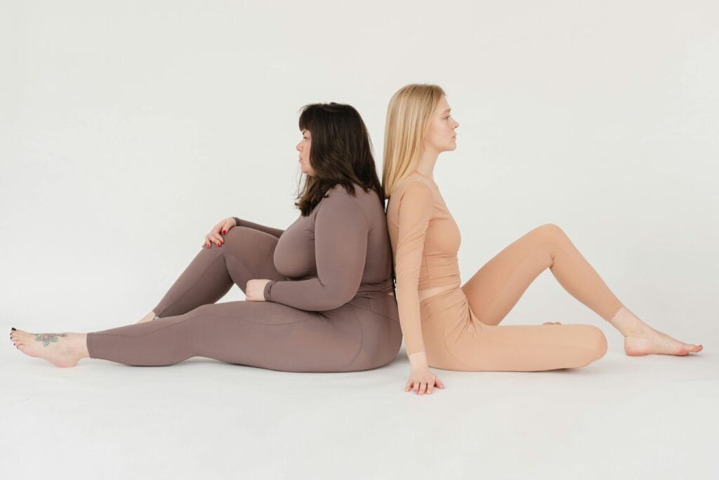 Two women of different body types sitting back to back on a white background, promoting body positivity.